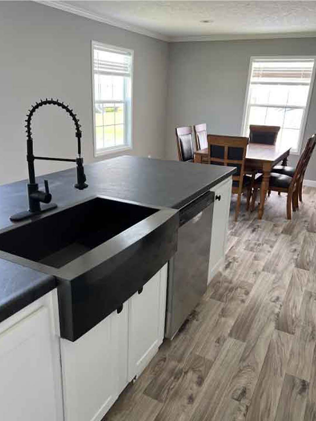 Modern kitchen with a black farmhouse sink and sleek faucet on a dark countertop. Light gray walls, large windows, and wooden dining table create a bright, cozy atmosphere.