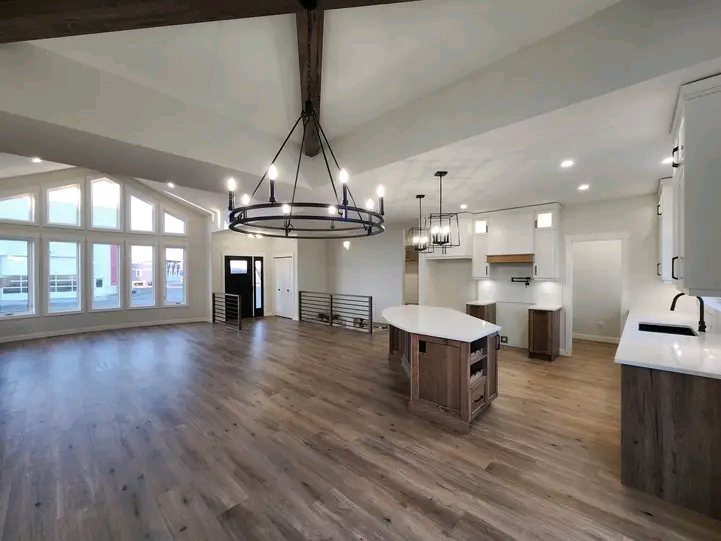 Spacious open-concept room with light wood floors, vaulted ceiling, and large arched windows. A modern kitchen features an island and elegant chandeliers.