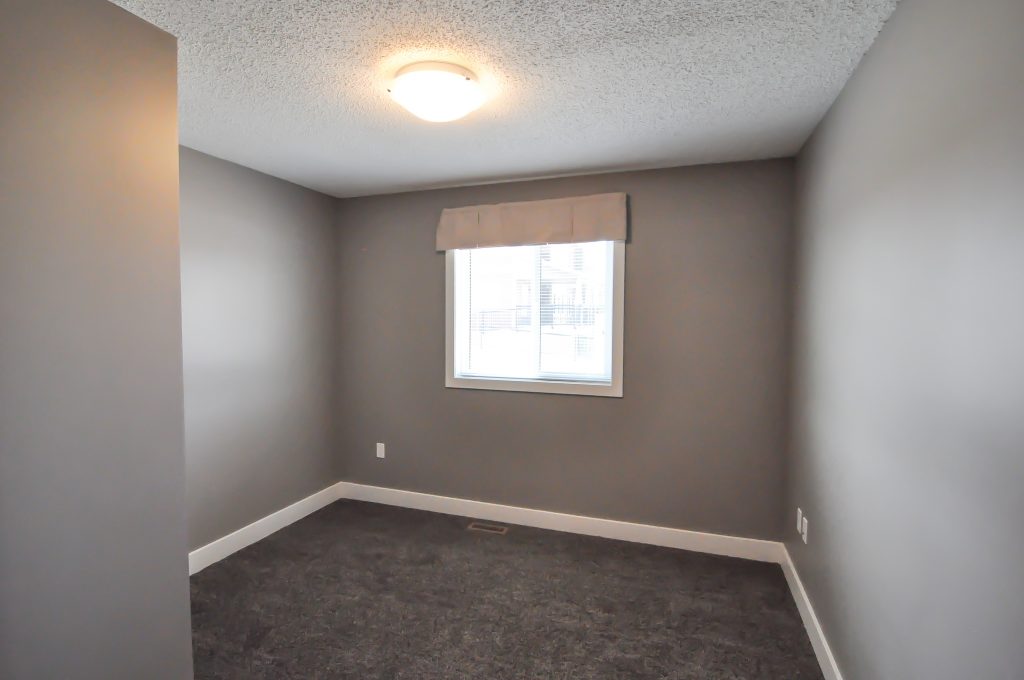 Small empty room with gray walls and carpet, a single window with a valance, and a ceiling light. The atmosphere is neutral and modern.