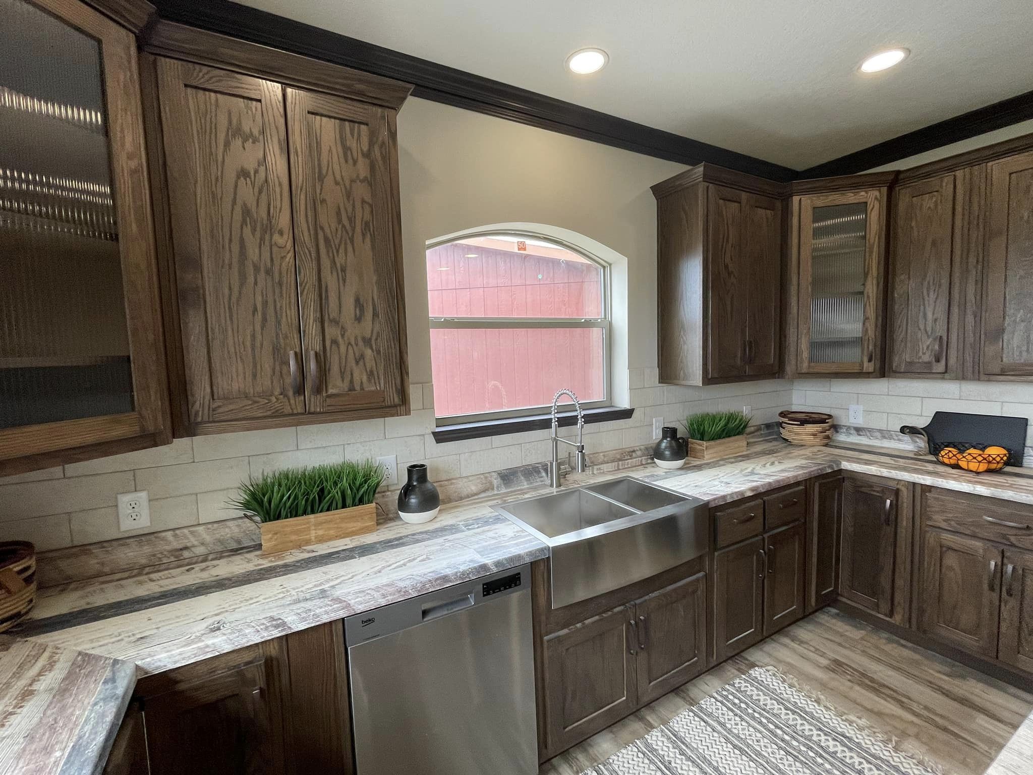 Modern kitchen with dark wood cabinets, a stainless steel sink under an arched window, light countertops, decorative plants, and a bowl of oranges. Cozy and inviting.