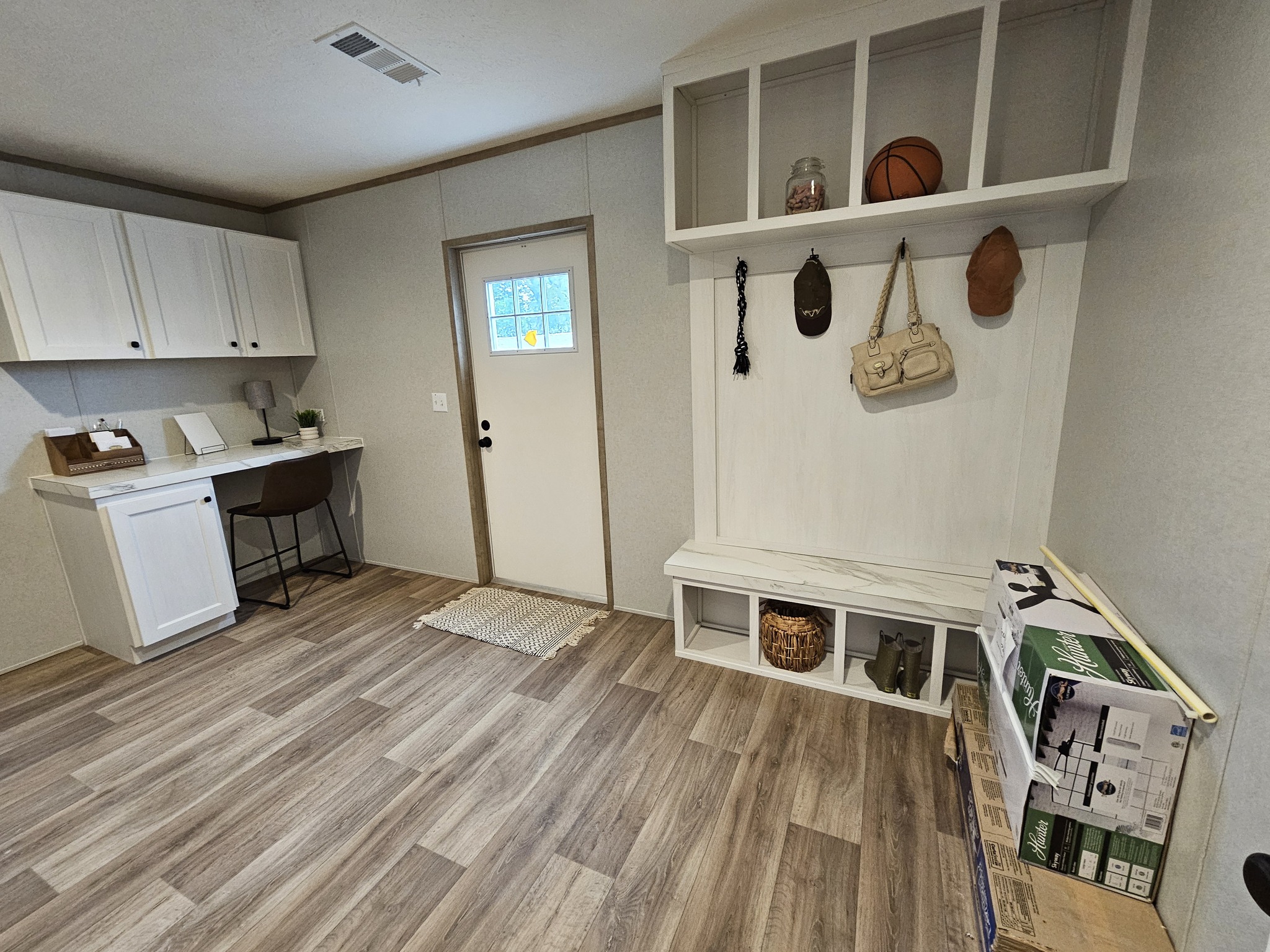 A cozy mudroom with a wooden bench and shelves holding a basketball and decor. There's a door with a small window, a home office corner, and boxes on the floor.
