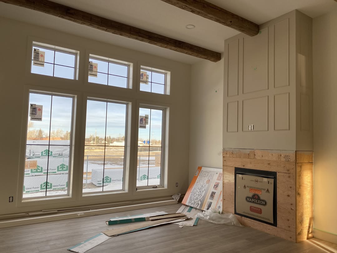Spacious room under construction, featuring large windows with natural light, exposed wooden beams, and an unfinished fireplace. The mood is warm and inviting.