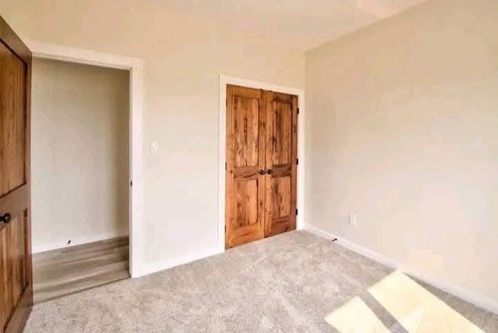 A small empty room with cream-colored walls and beige carpet. Wooden double doors and a simple wooden door lead to other areas. Sunlight creates a warm, welcoming glow.