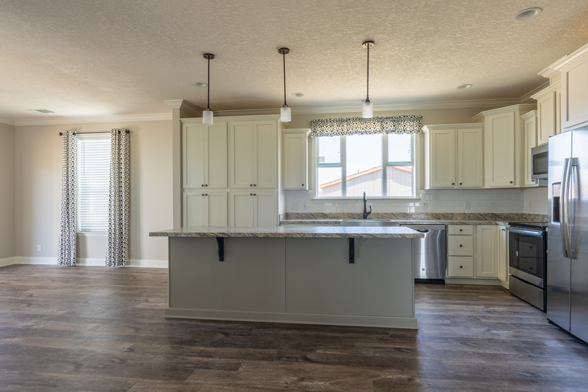 Modern kitchen with white cabinets, stainless steel appliances, and a granite island. Three pendant lights hang above, creating a spacious, elegant feel.