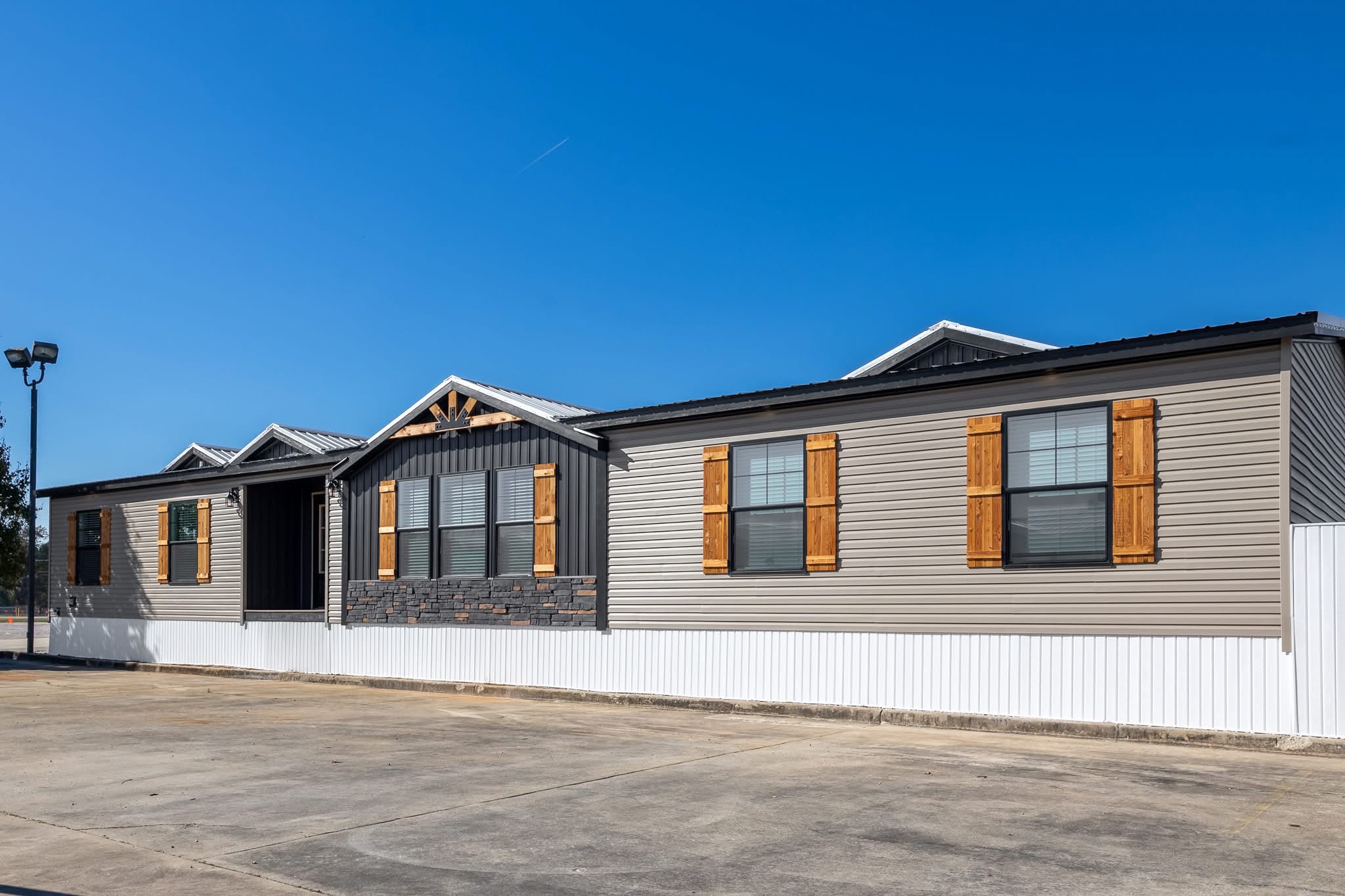 A modern manufactured home with tan siding, wooden shutters, and a section of stone accent under a clear blue sky. The driveway is empty, conveying tranquility.