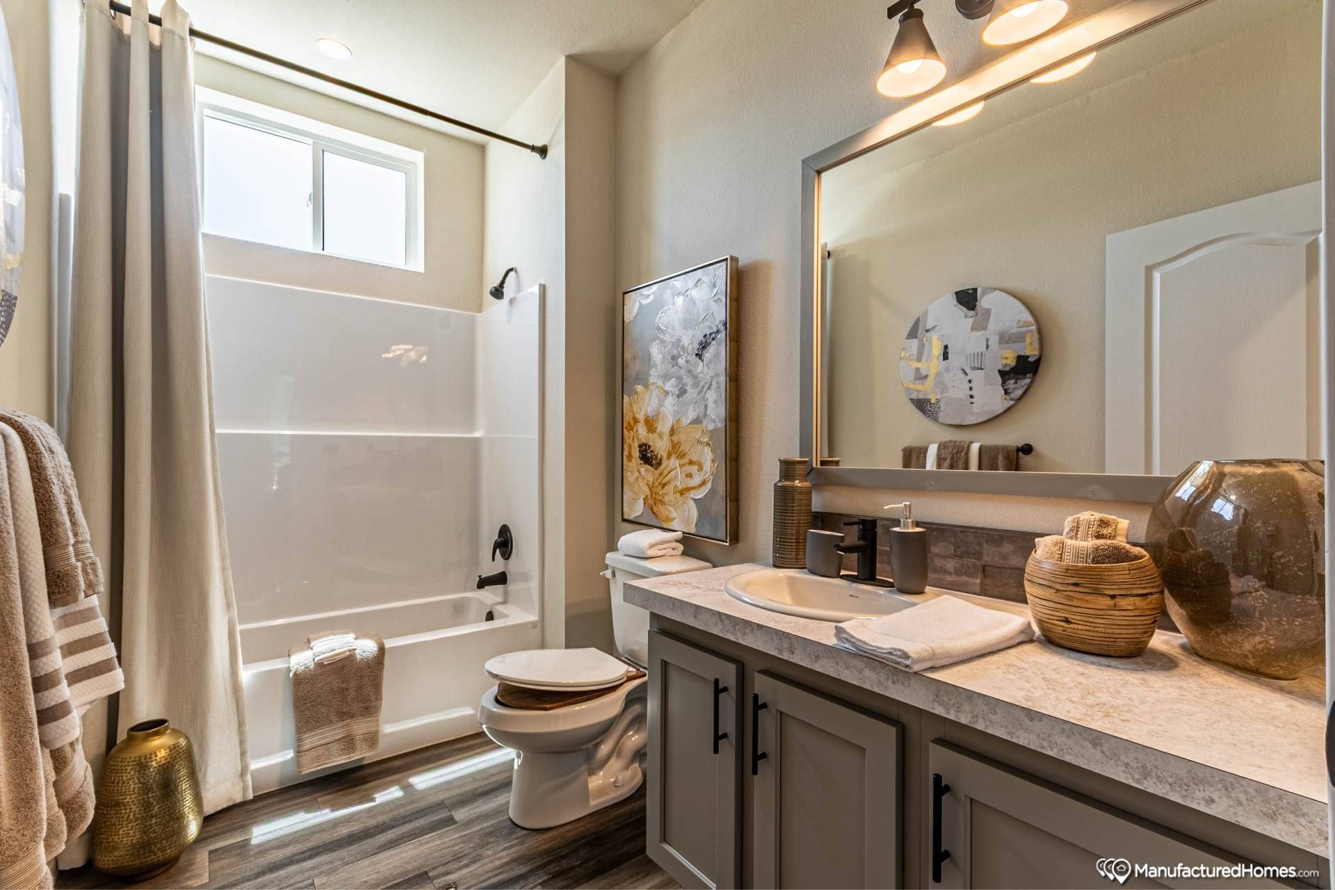 A modern bathroom with a white tub and shower, light grey cabinets, and a large mirror. Decor includes abstract art and pottery. Bright and cozy ambiance.