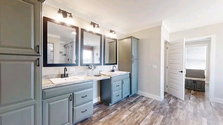 Modern bathroom featuring dual sinks with marble countertops, sleek gray cabinets, large mirrors, wood flooring, soft lighting, and an open door to another room.