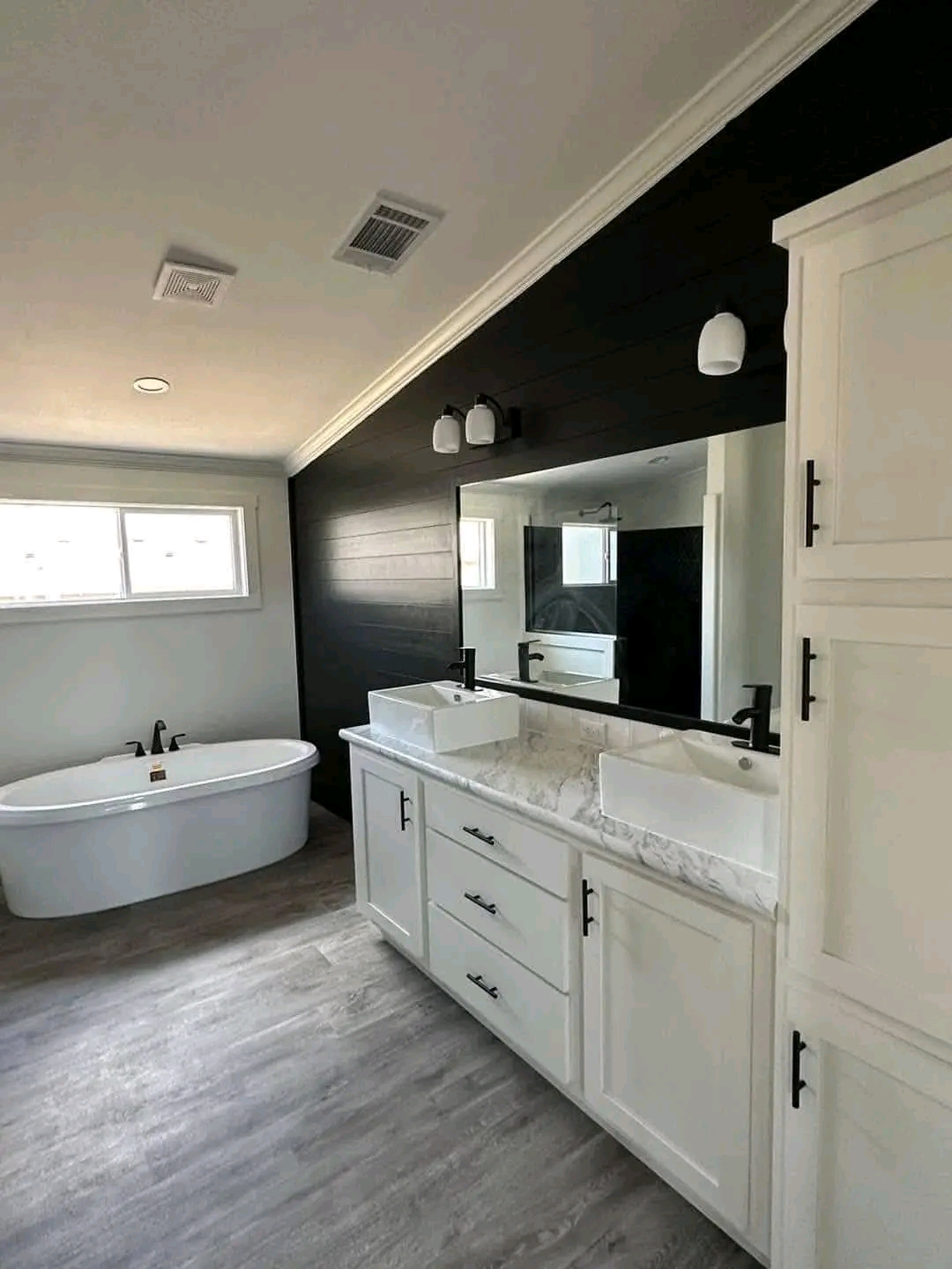 Modern bathroom with a sleek white double vanity and marble countertop, lined with black fixtures. A freestanding bathtub sits by a large window, enhanced by a black accent wall for a chic, serene ambiance.