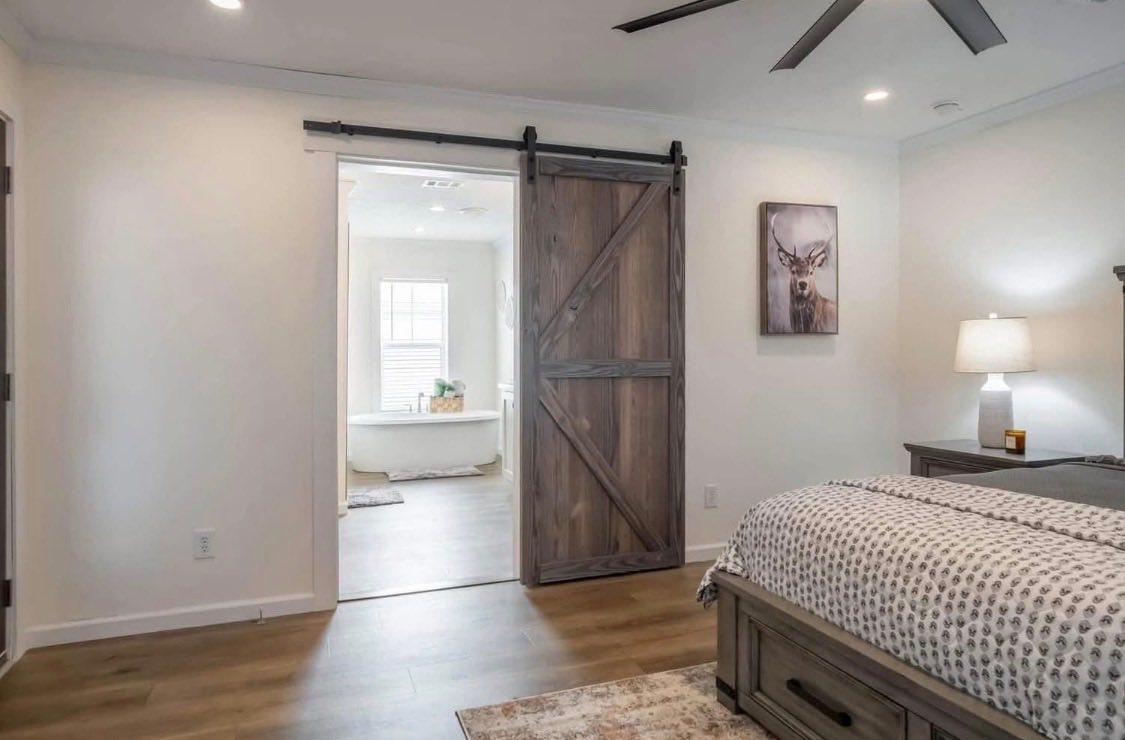 A cozy bedroom with a wooden sliding barn door leading to a well-lit bathroom. Features include a patterned bedspread, a nightstand with a lamp, and a deer artwork. The atmosphere is calm and inviting.