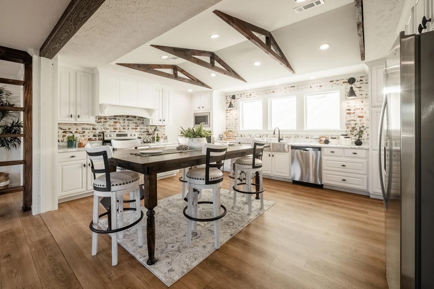 Stylish kitchen with wooden floor, exposed beams, island table with high chairs, white cabinets, brick backsplash, and large windows. Bright and welcoming.