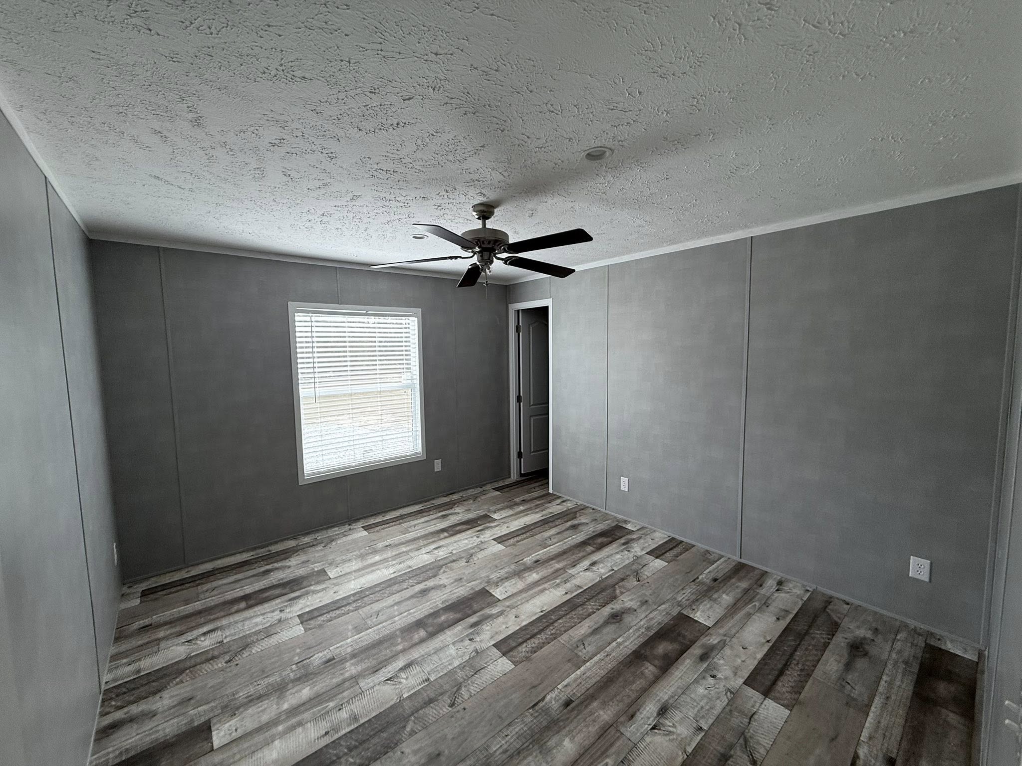 Empty room with gray walls and weathered wood floor, featuring a ceiling fan and a window with blinds. There's a closed door, creating a serene, minimalistic atmosphere.