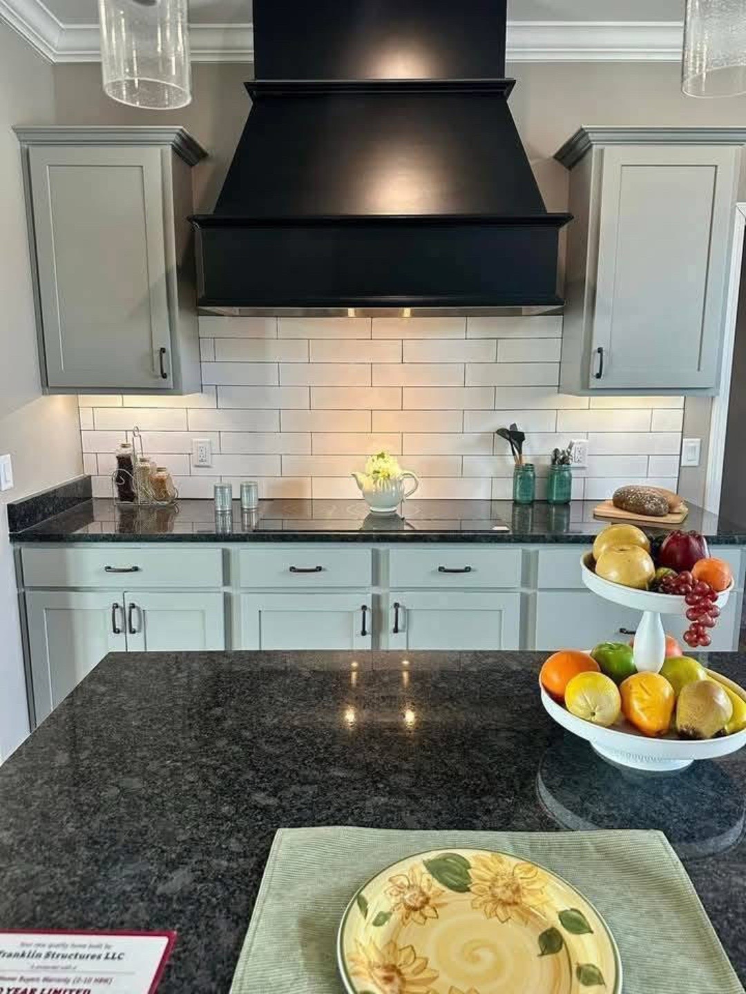 Modern kitchen with a black range hood, white subway tile backsplash, and gray cabinets. A fruit bowl and floral plate add color to the dark countertop.