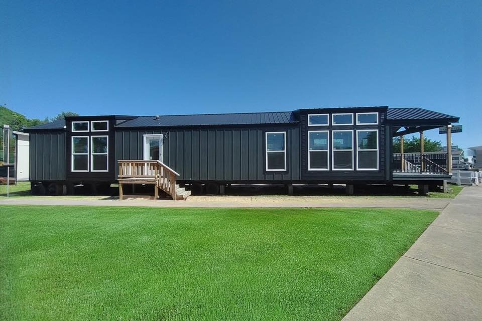 Modern black tiny house with large windows, wooden steps, and a veranda. Set on fresh green grass under a clear blue sky, conveying a calm, minimalist vibe.