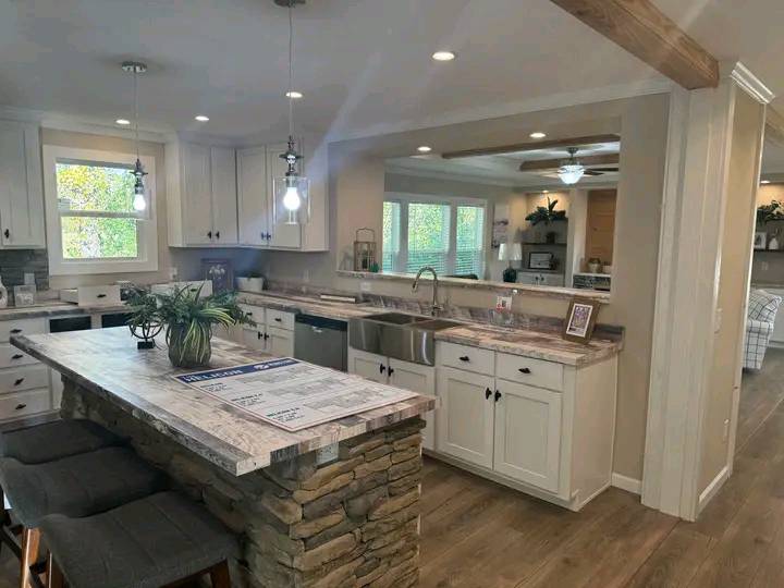 Bright kitchen with white cabinets, stone island, and wooden floors. Pendant lights hang above the island, and a cozy living room is visible in the background.