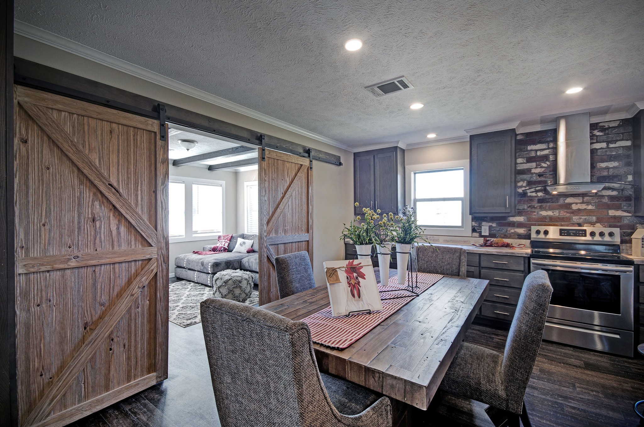 Cozy farmhouse kitchen with rustic wooden sliding barn doors. A wooden dining table with red checkered runner hosts flowers, creating a welcoming vibe.