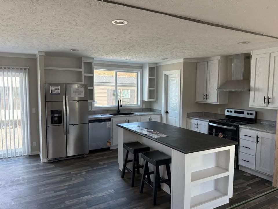 Modern kitchen with white cabinets, gray countertops, and a central island with stools. Stainless steel appliances and large window create a bright, airy feel.