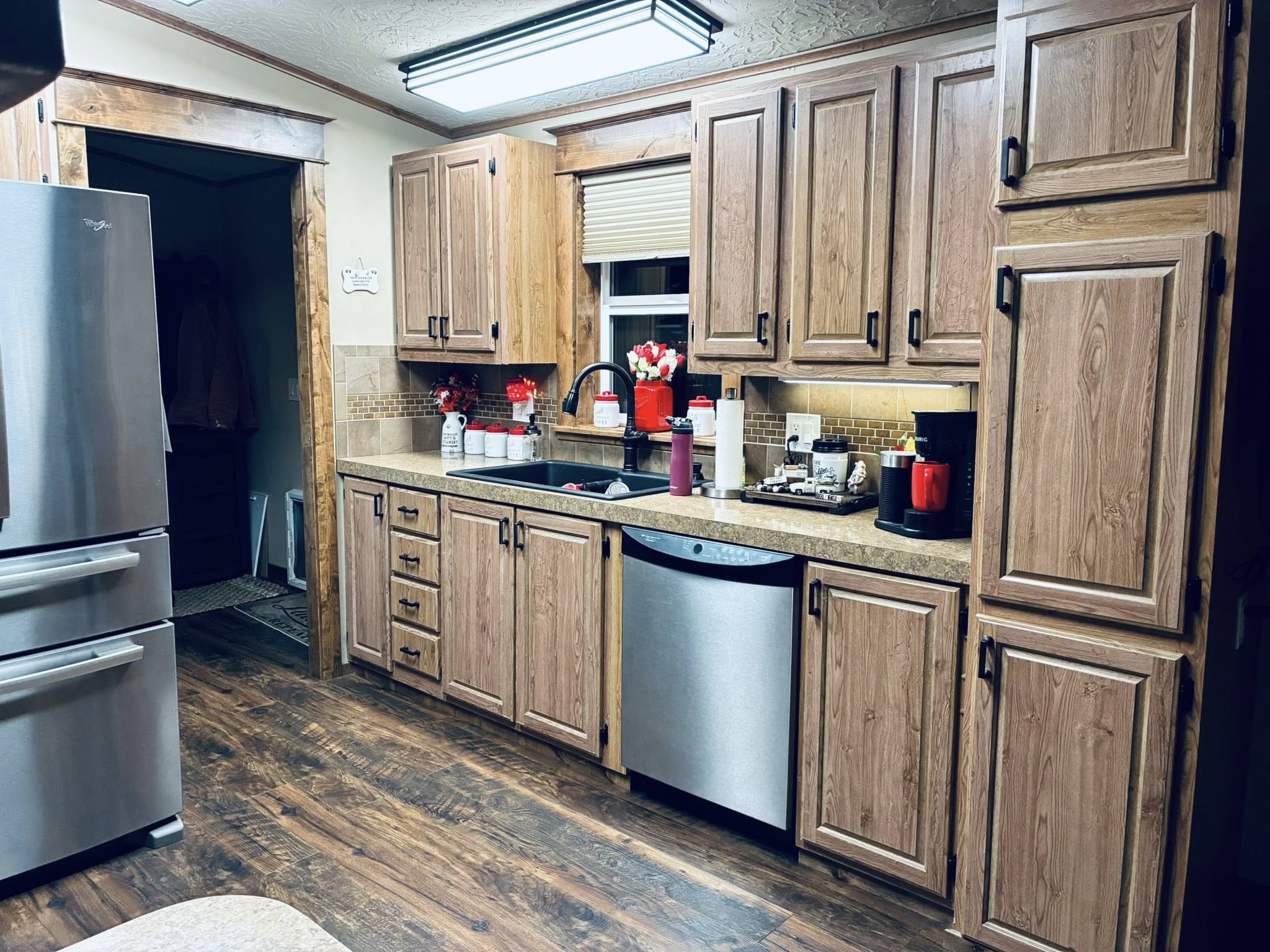 Cozy kitchen with wooden cabinets, stainless steel appliances, and warm lighting. Red accents include a kettle and flower vase by the window, enhancing the inviting atmosphere.