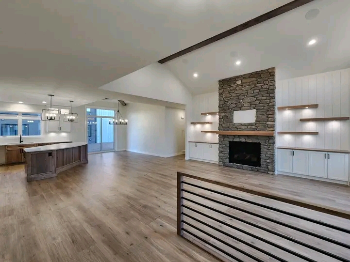 Spacious modern living room with vaulted ceilings, a stone fireplace, and open kitchen. Wood floors, minimalistic decor, and warm lighting create a cozy ambiance.