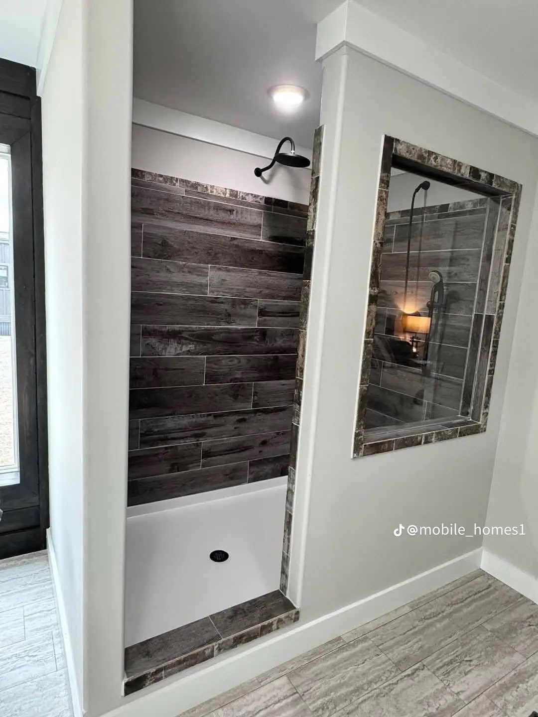 Modern open shower with dark wood-like tiles and a sleek black showerhead. Adjacent is a large mirror framed in similar tiles, adding elegance.
