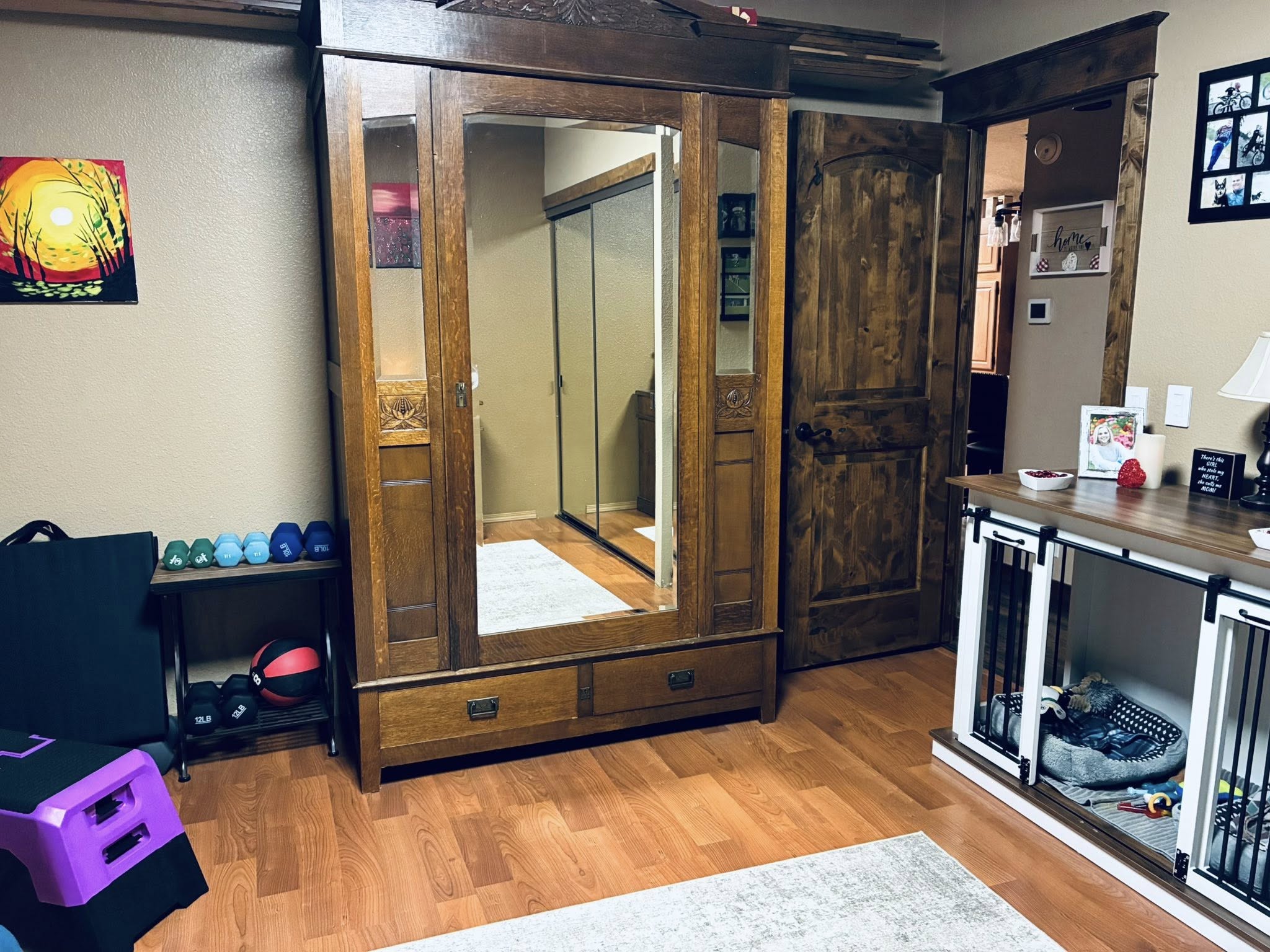 Cozy room with wooden floor, featuring a large wooden wardrobe with a mirror, a small fitness area with weights, and a console table with decor.