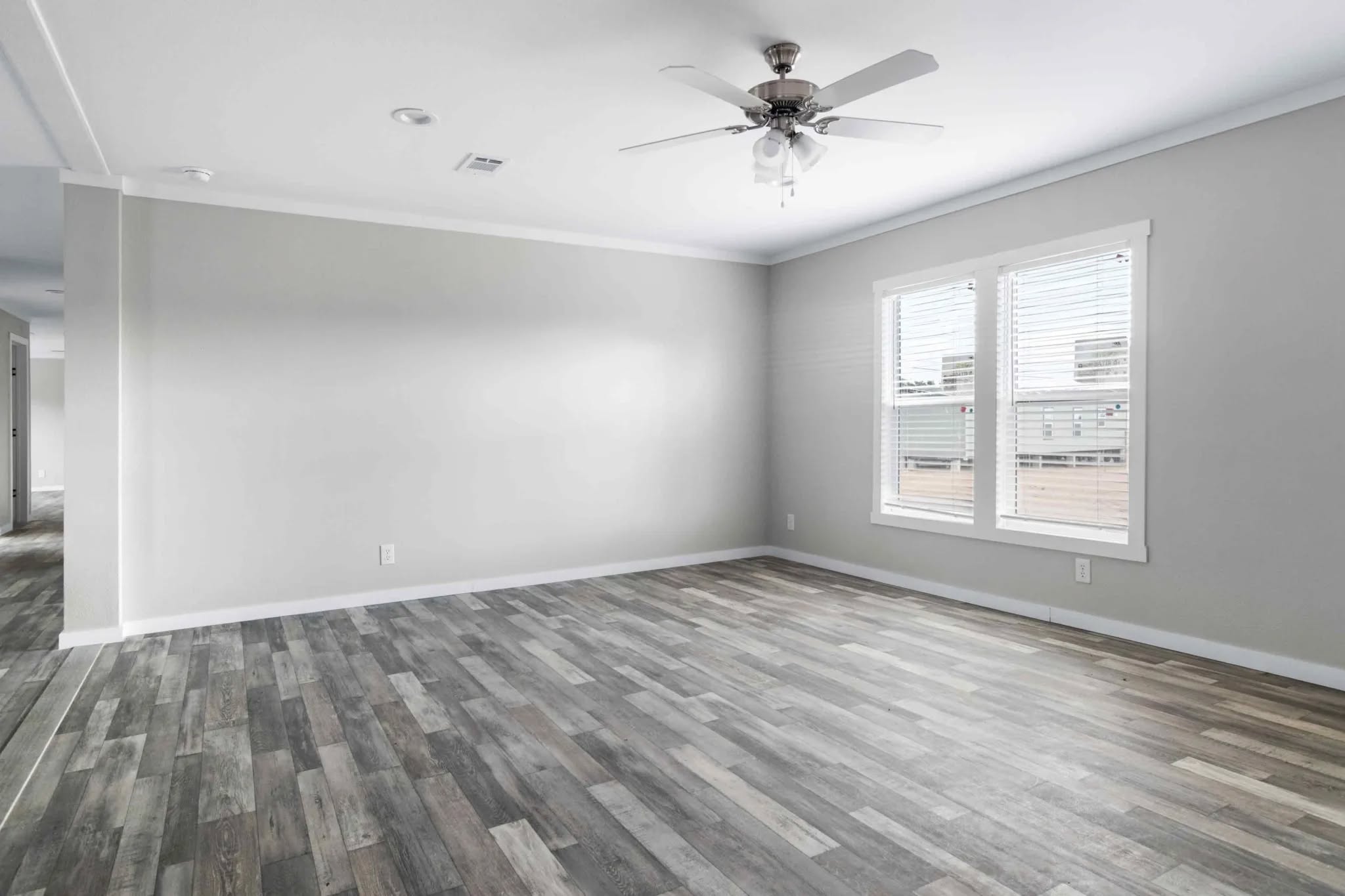 Empty room with light gray walls, wood-style flooring, and two windows with white blinds. Ceiling fan overhead, creating a bright, spacious feel.