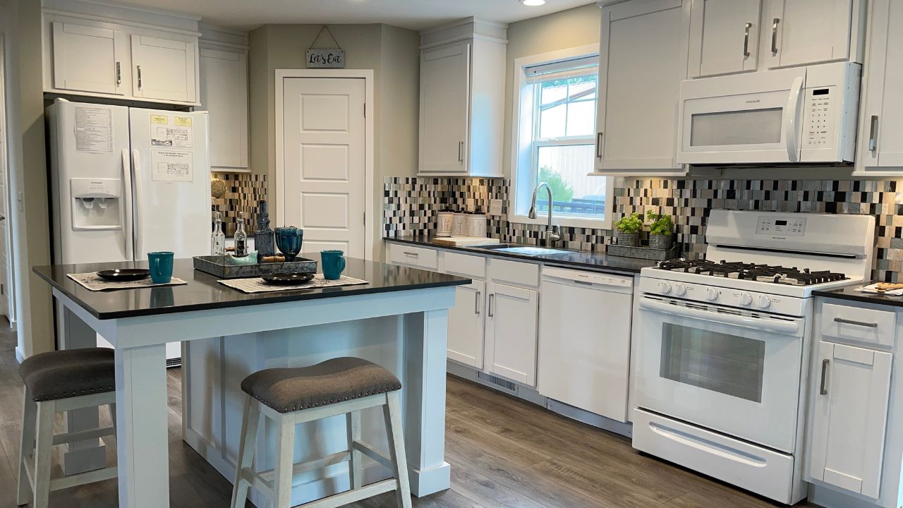 A modern kitchen with white cabinets, a tiled backsplash, and wood flooring. Features include a central island with stools, a white fridge, oven, and a window.