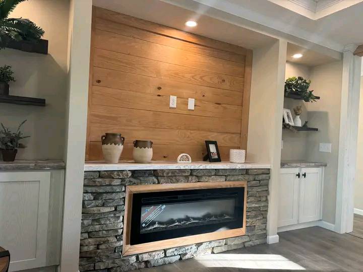 Modern living room with a stone-framed electric fireplace, wooden accent wall, decorative vases, shelves with plants, and soft overhead lighting.