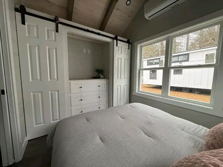 Cozy bedroom with a gray bed, barn-style closet doors revealing white drawers. Large window shows a tiny house outside. Warm, rustic ambiance.