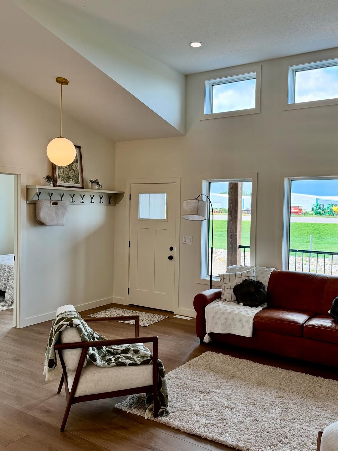 A cozy living room with a vaulted ceiling, featuring a red sofa, a modern chair with a green throw, light wood flooring, and a large rug. The space has a neutral palette, a round pendant light, and a shelf with hooks above the entrance. Large windows offer a view of greenery outside.