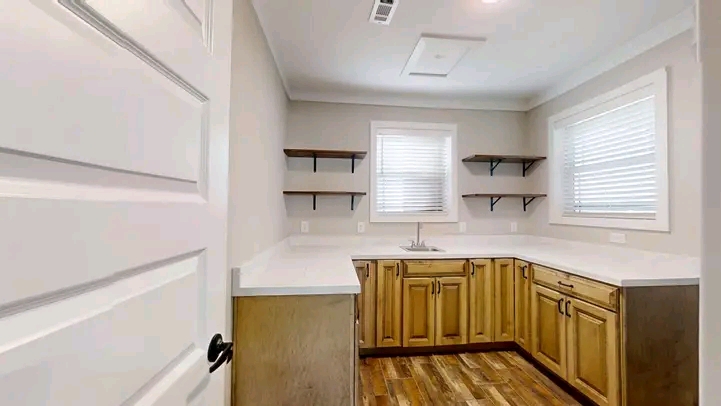 Bright laundry room with wooden cabinets, white countertops, and open shelves. Two windows let in natural light. Hardwood floor adds warmth.