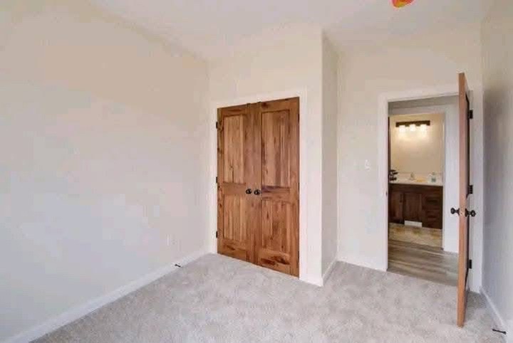 Empty room with beige walls and carpet, featuring a wooden closet door and an open door leading to a lit bathroom, creating a calm atmosphere.