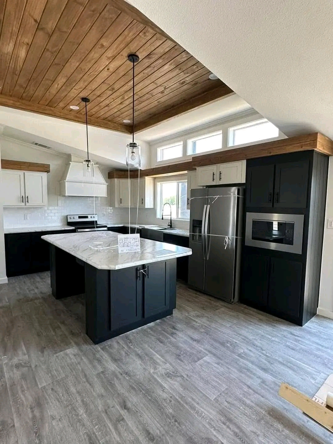 Modern kitchen with a wood-paneled ceiling, dark cabinetry, white countertops, stainless steel appliances, and pendant lights. Bright, inviting space.