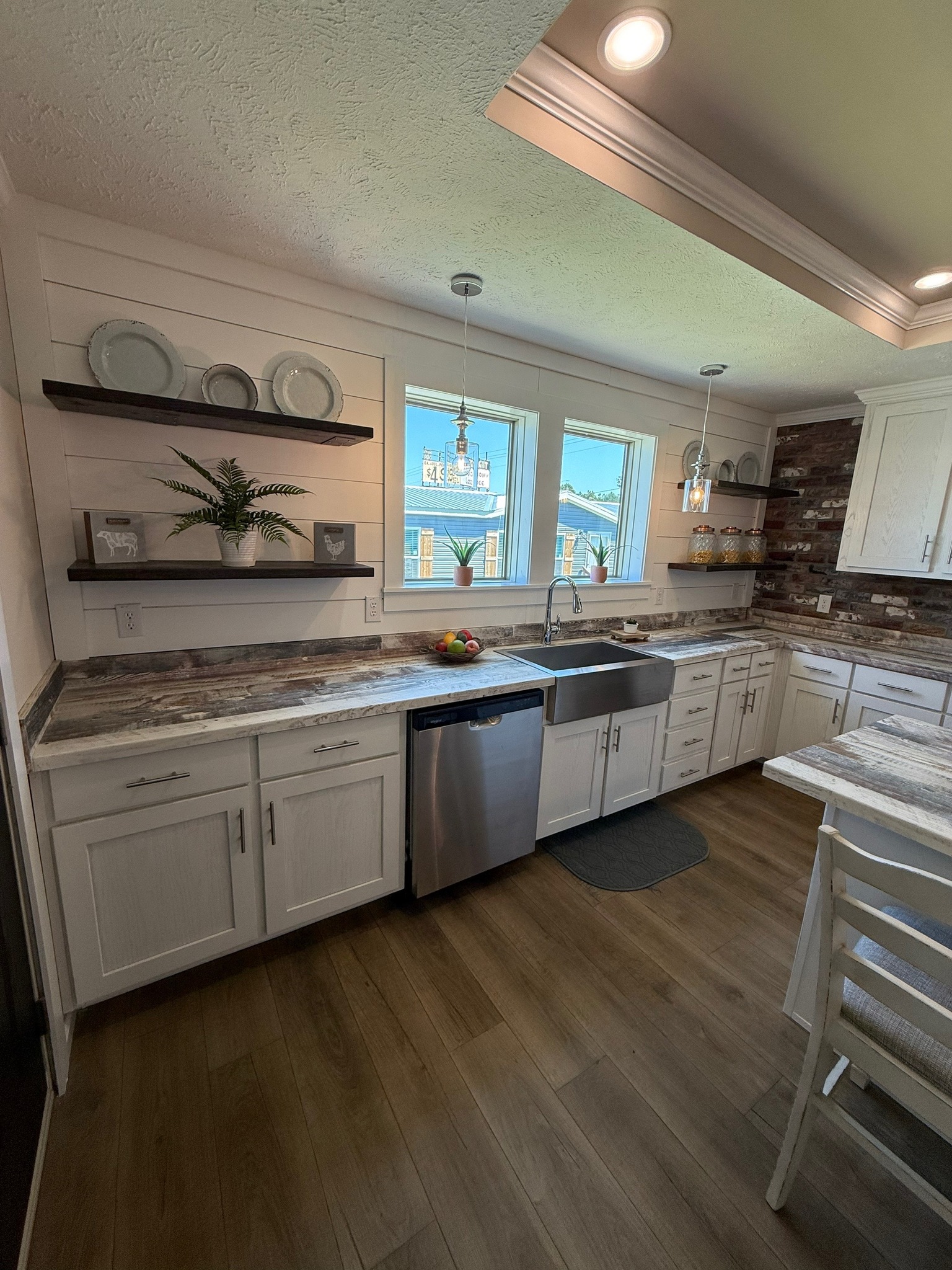 Modern kitchen with white cabinets, wooden floors, and stone countertops. Sunlight streams through two windows, highlighting the farmhouse sink and open shelves with dishes and a plant. The atmosphere is bright and inviting.