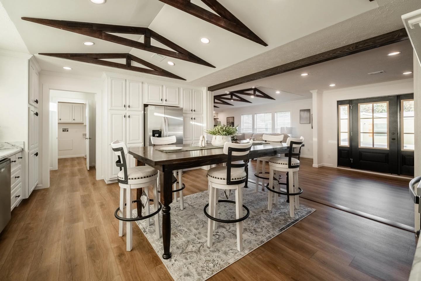 Spacious kitchen with wood floors, a central dark wood island, and high chairs. White cabinets, stainless steel fridge, and rustic ceiling beams.