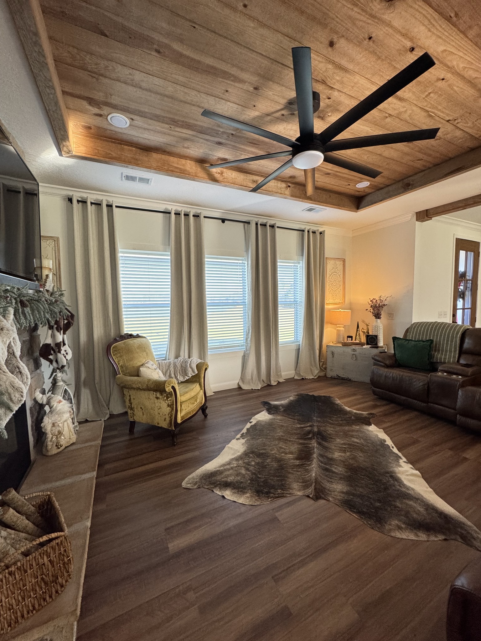 Warm living room with wood ceiling, large fan, and cozy decor. Features a brown leather sofa, vintage armchair, cowhide rug, and soft lighting.