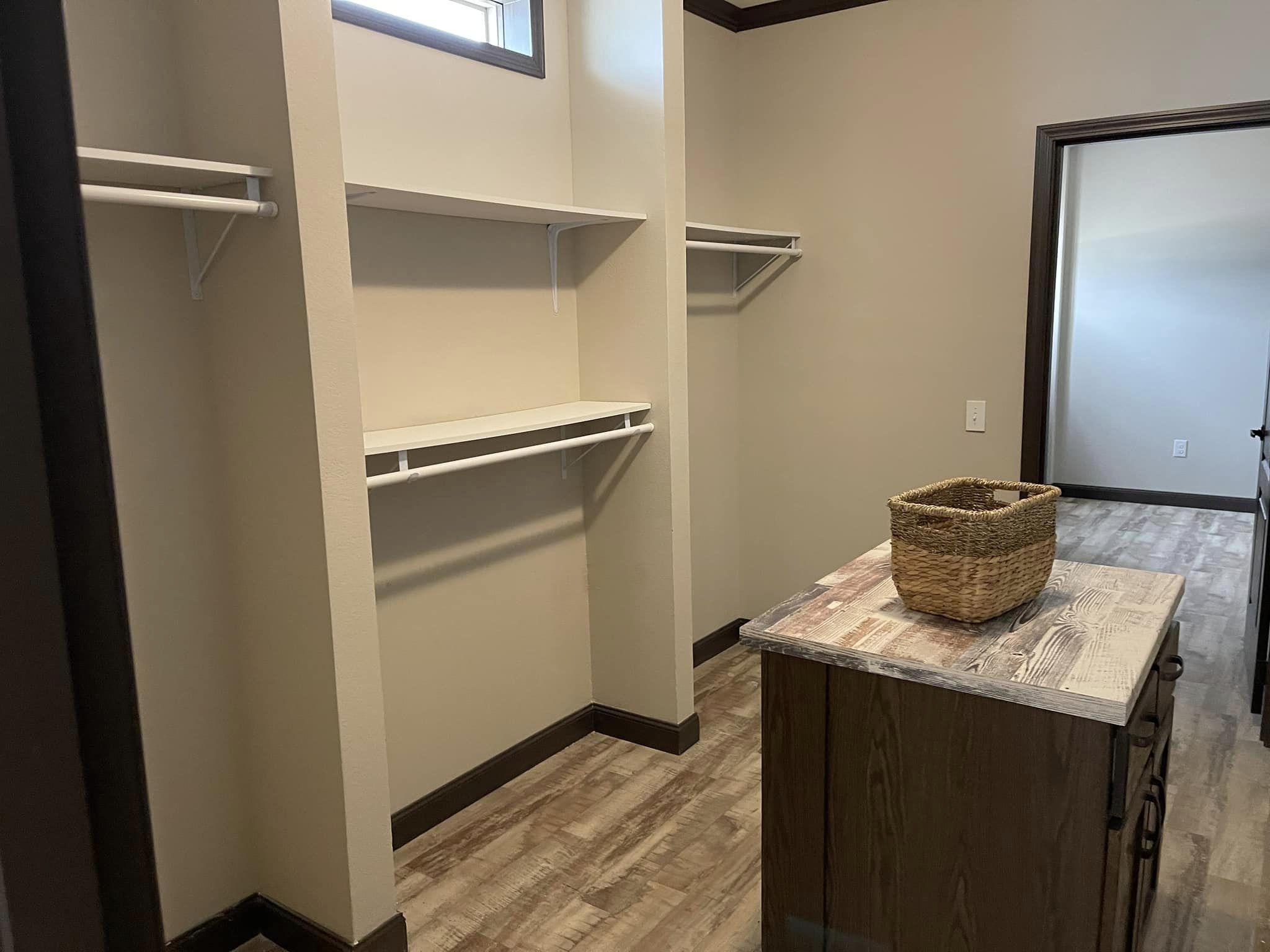 A spacious walk-in closet with light walls features wooden floors, open shelves, and hanging rods. A basket sits on a central wooden island.