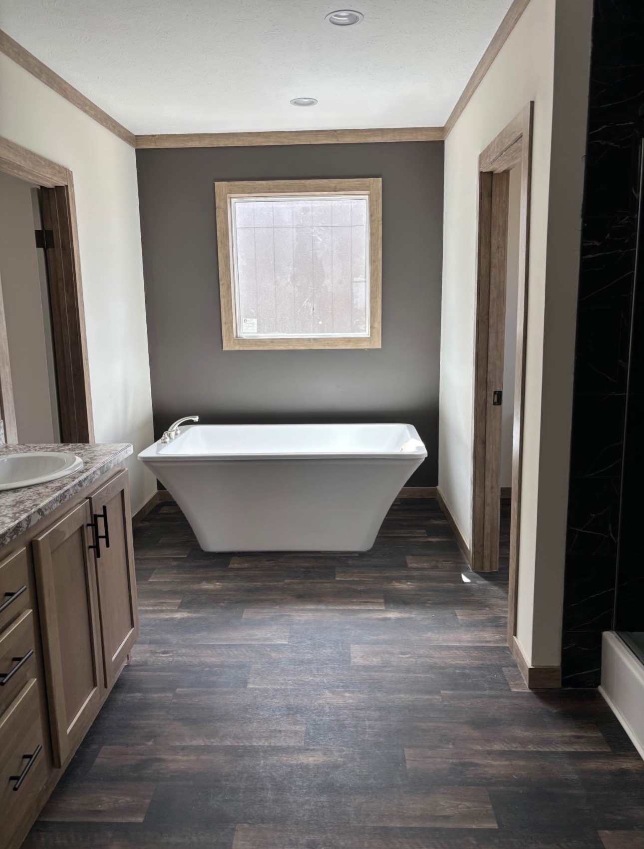 Modern bathroom with a freestanding white tub beneath a window on a dark accent wall. Wood cabinets and flooring create a warm, inviting tone.