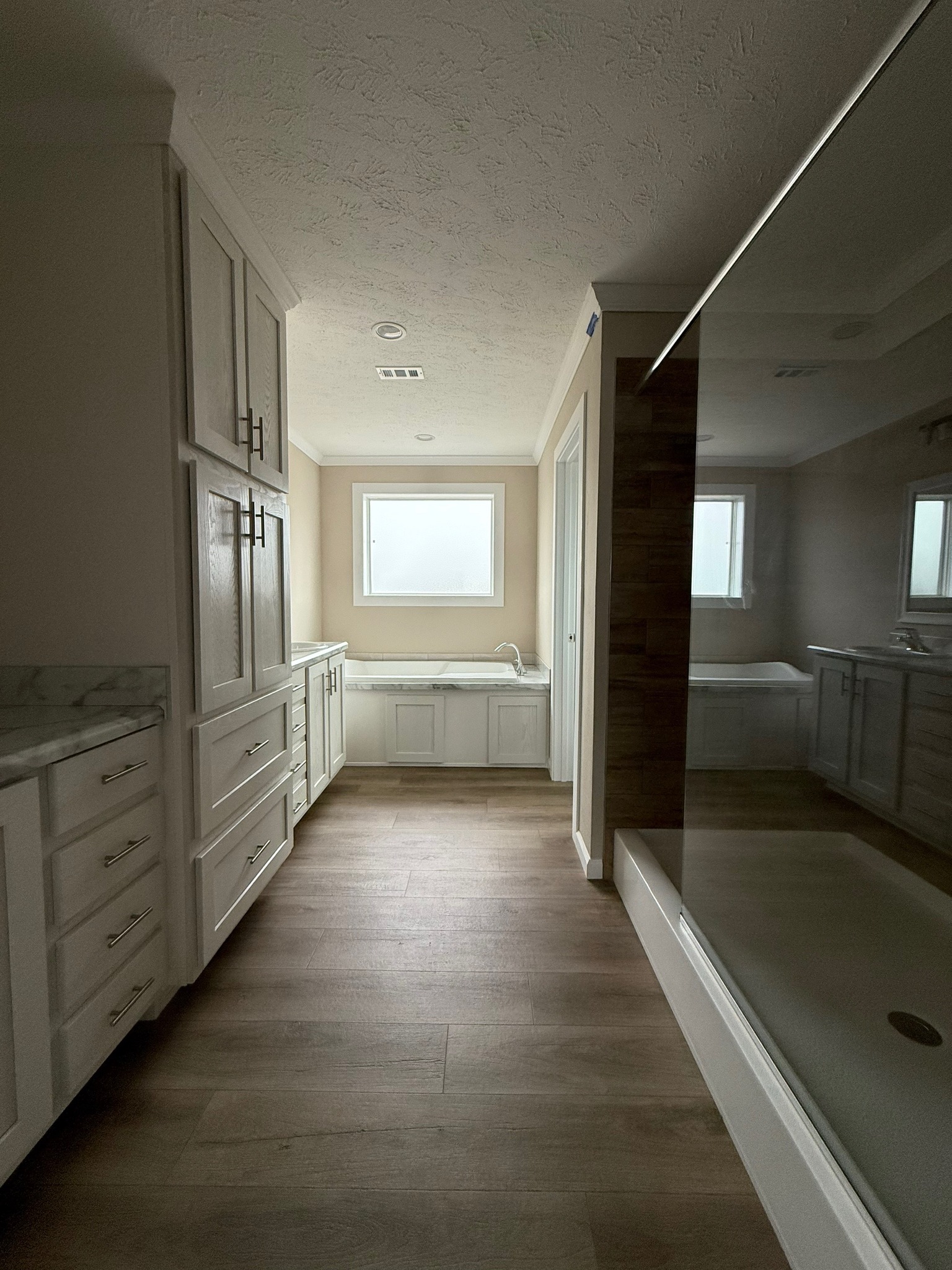 Modern bathroom with a large walk-in shower on the right, white cabinets and a marble countertop on the left, and a window above a bathtub in the background.