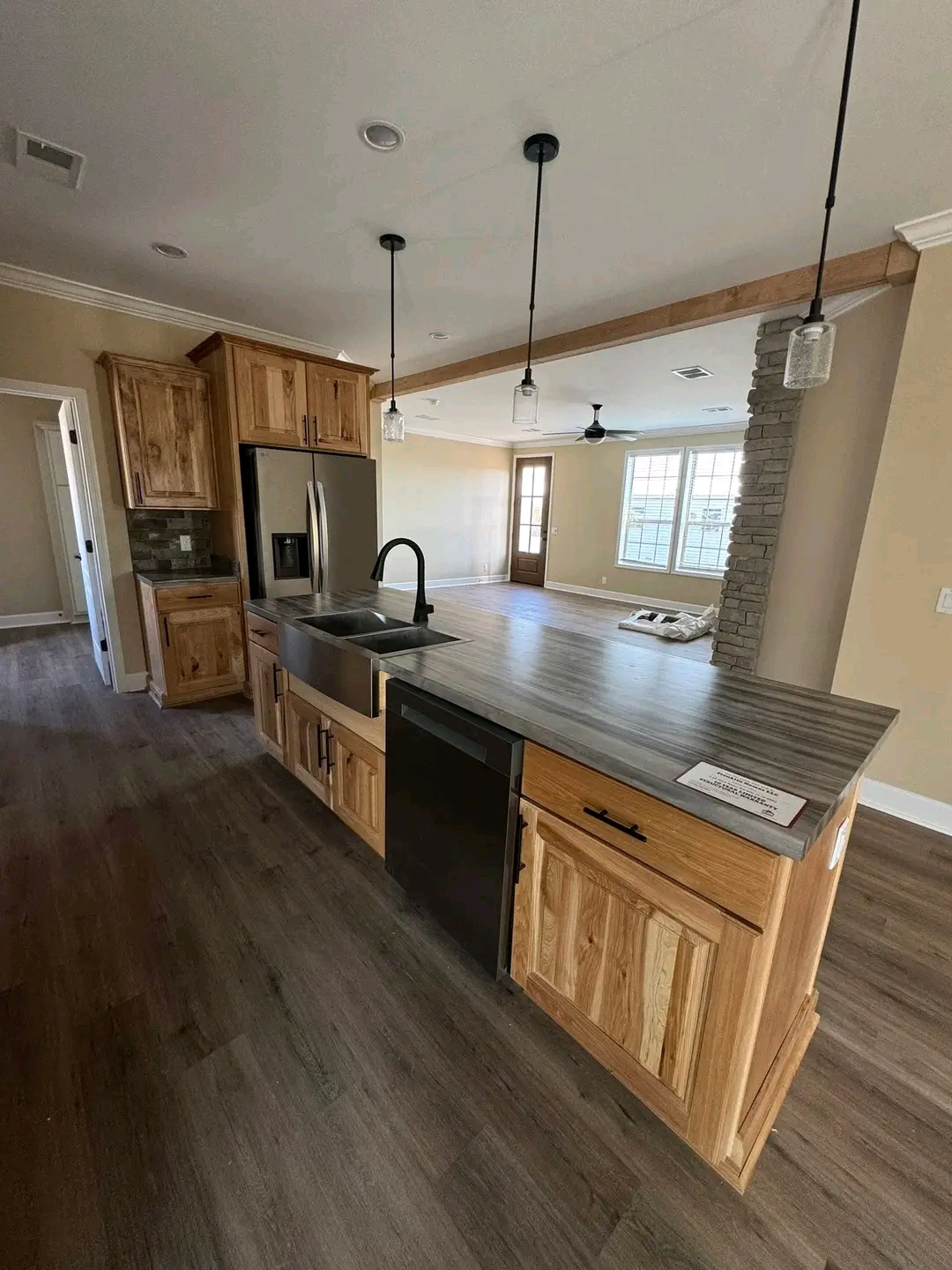 Modern kitchen with warm wood cabinets and a gray countertop. Features a stainless steel fridge, farmhouse sink, pendant lights, and open view to living area. Cozy and inviting.