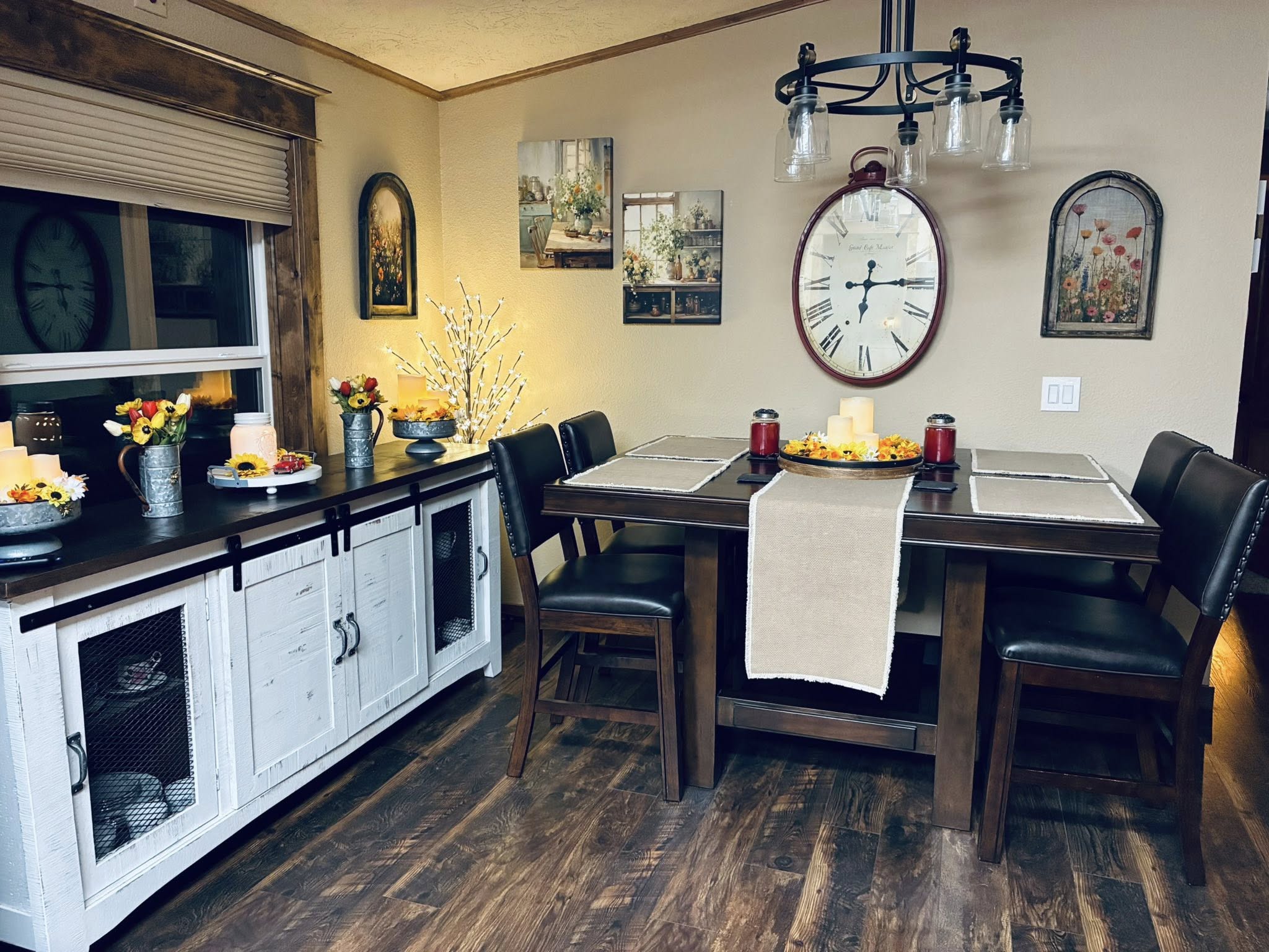 Cozy dining room with rustic charm. Wooden table set with beige runners, surrounded by black chairs. Large wall clock and floral art on cream walls. White sideboard with candles and small bouquets creates a warm, inviting atmosphere.