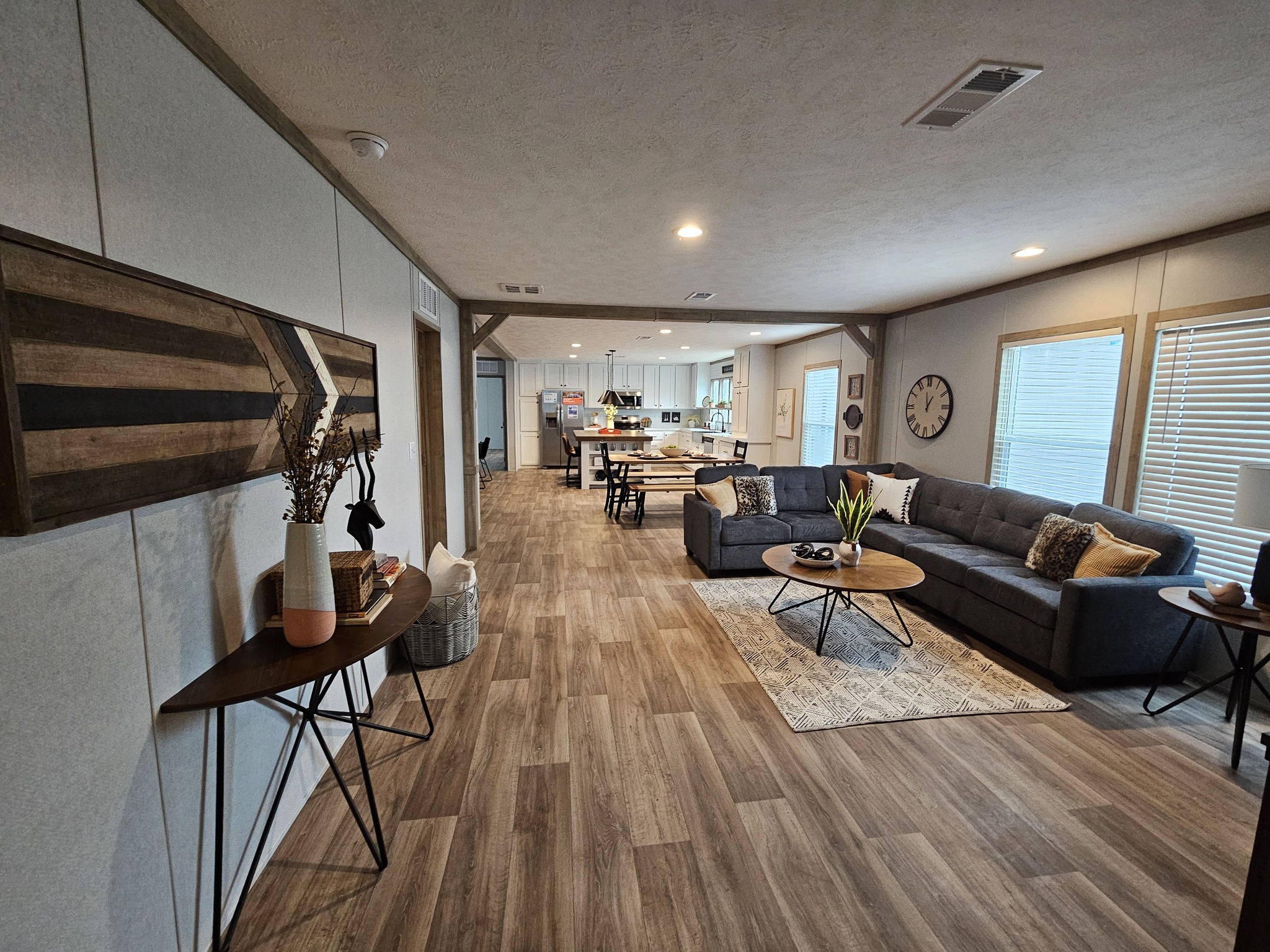 Spacious living room with wood flooring and gray sectional sofa, decorated with pillows. Adjacent kitchen and dining area, modern, bright, and welcoming.
