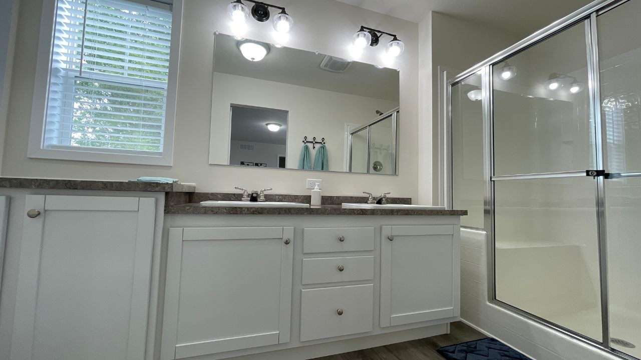 A modern bathroom with white cabinets, dual sinks, large mirror, and bright lighting. Glass shower enclosure on the right and a window with blinds on the left.