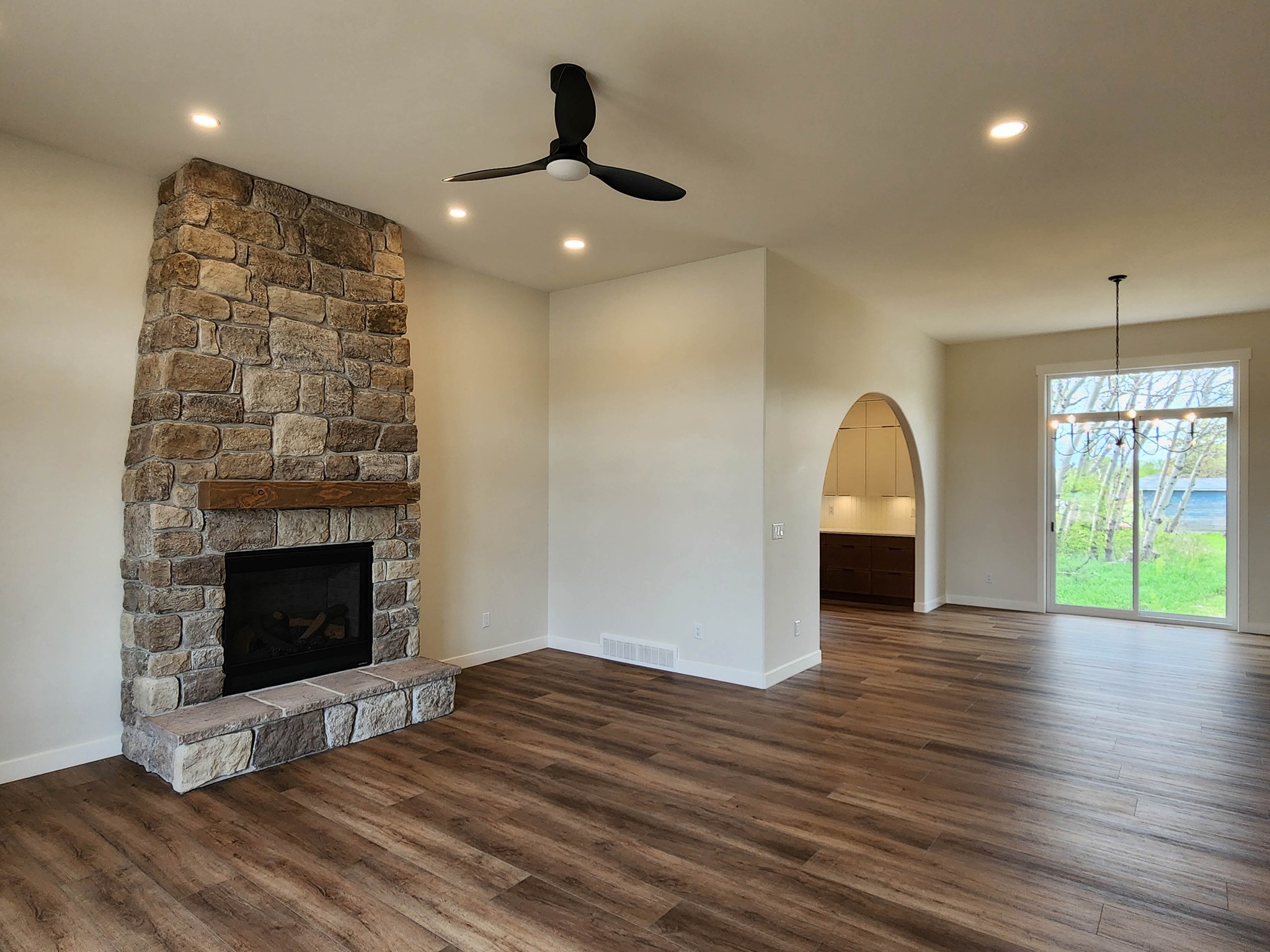 A modern living room with a stone fireplace on the left, wood floors, a ceiling fan, and wide patio doors on the right revealing a lush green view.
