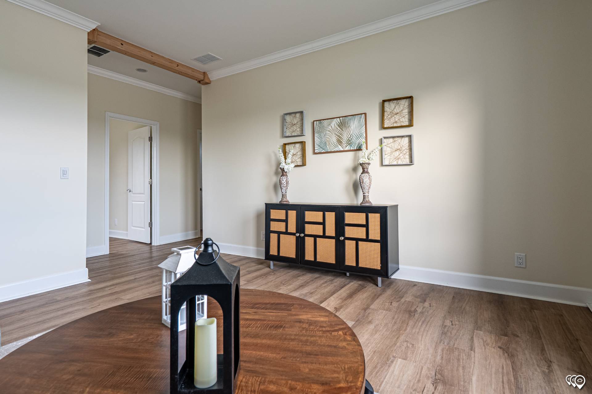 Minimalist living room with light wood flooring and beige walls. A black cabinet with wicker accents is adorned by tall vases. Framed leaf artwork hangs above.