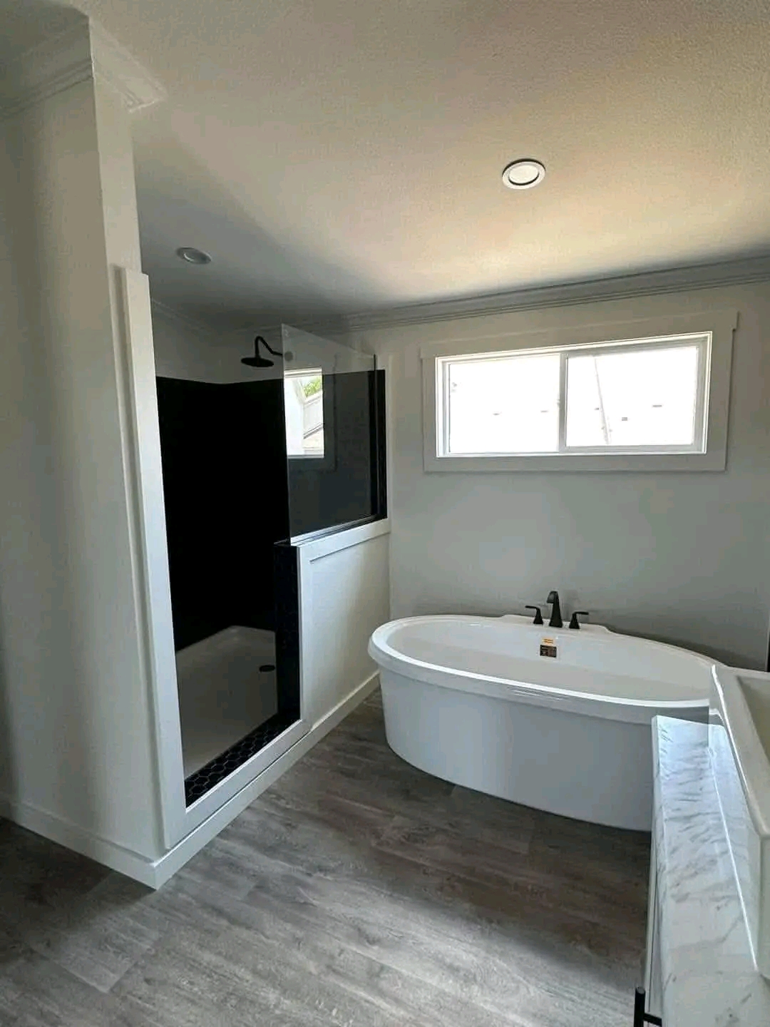 Modern bathroom with a white freestanding bathtub under a window, a glass-enclosed shower with dark tiles, and wood-effect flooring. Bright and minimalist.