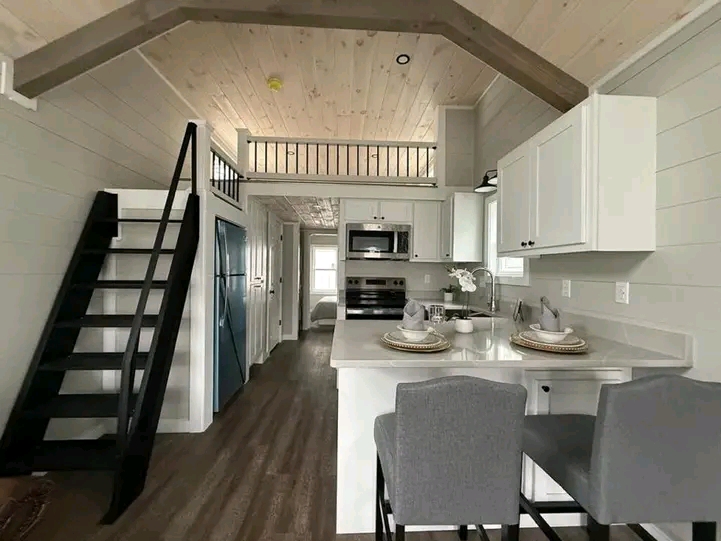 Cozy kitchen in a tiny house with white cabinets, a breakfast bar set for two, grey chairs, a black metal loft ladder, and wood accent ceiling.