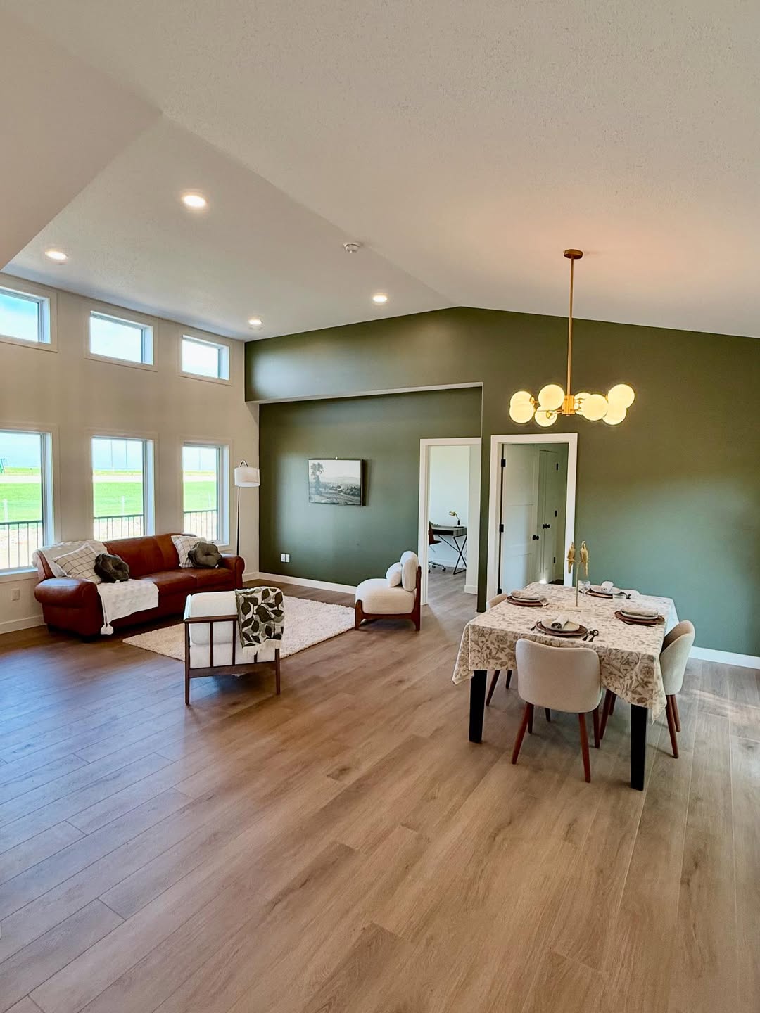 Spacious living-dining area with wooden floors and green walls. Modern chandelier, elegant furniture, and large windows with natural light create a cozy ambiance.