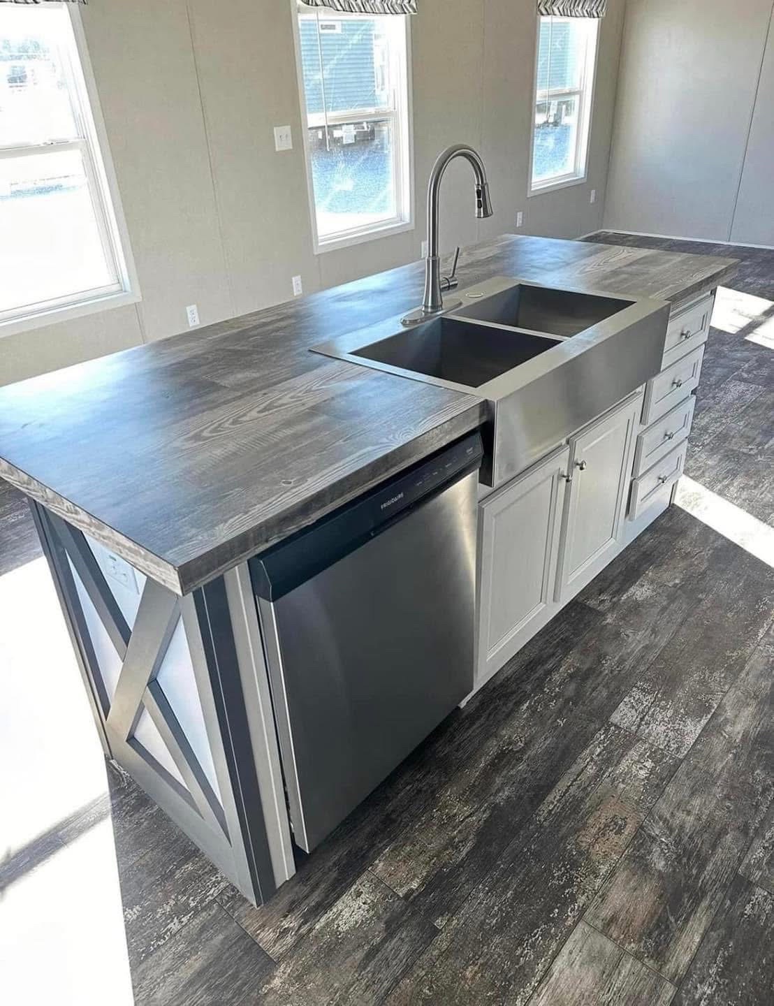 Modern kitchen island with a sleek gray countertop, stainless steel sink, dishwasher, and white cabinets. Sunlight streams through three windows.
