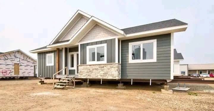 A modern house with gray siding, white trim, and a stone accent wall sits elevated on blocks. Steps lead to the front porch, with a clear sky overhead.