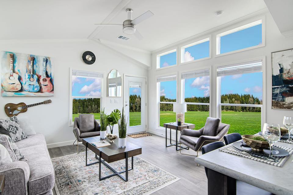 Modern living room with large windows showcasing a scenic green landscape. Features gray sofas, a guitar on the wall, and vibrant artwork, creating a bright and inviting atmosphere.